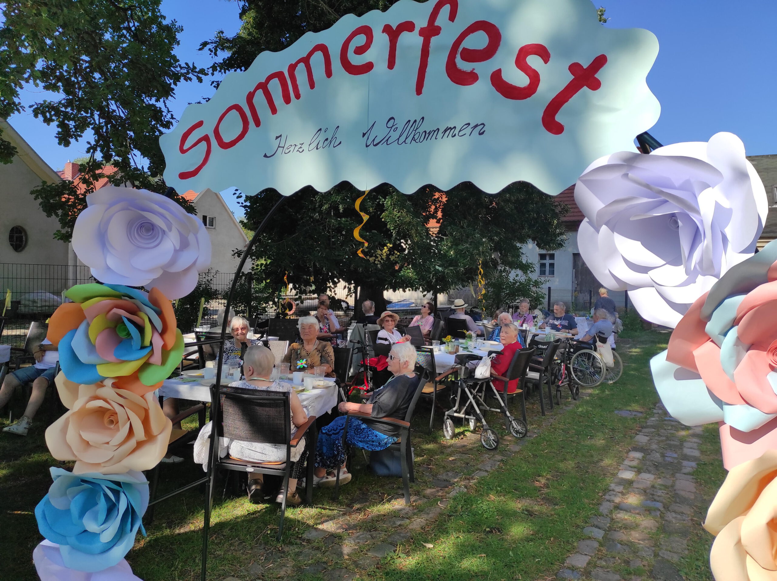 Gäste sitzen im Garten an Tischen. vorne steht ein Schild mit der Aufschrift Sommerfest Herzlich Willkommen